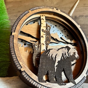 May include: A wooden wall art piece featuring a layered design of a black bear and two cubs. The bear is standing on a snowy mountain scene with a birch tree in the background. The piece is made of wood and has a rustic look.