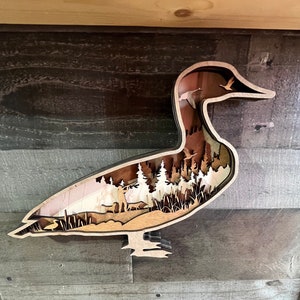 May include: A wooden cutout of a goose with a forest scene inside. The goose is brown and the forest scene is made of white wood.