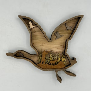 May include: A wooden wall art piece depicting a flying goose with a hunter and dog in the field below. The goose is made of layered wood with a cutout design. The hunter and dog are also made of wood and are silhouetted against the field.
