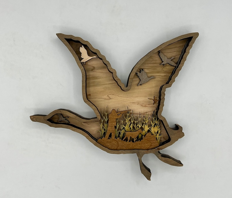 May include: A wooden wall art piece depicting a flying goose with a hunter and dog in the field below. The goose is made of layered wood with a cutout design. The hunter and dog are also made of wood and are silhouetted against the field.