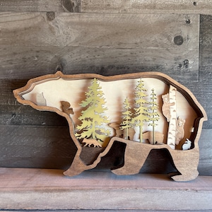 K&ouml;nnte beinhalten: Wanddekoration aus Holz in Form einer B&auml;ren-Silhouette mit einem Ausschnitt, der eine Waldszene mit B&auml;umen, Tieren und einer Birke zeigt.