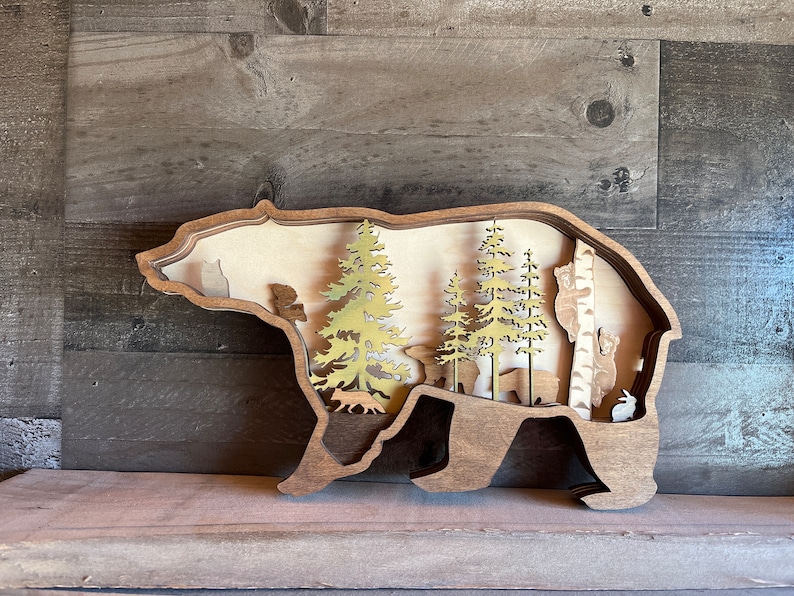 K&ouml;nnte beinhalten: Wanddekoration aus Holz in Form einer B&auml;ren-Silhouette mit einem Ausschnitt, der eine Waldszene mit B&auml;umen, Tieren und einer Birke zeigt.