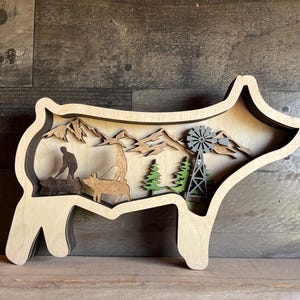 May include: A wooden cutout of a pig with a layered design. The pig is standing on a wooden surface. The cutout features a scene of a person walking a dog in a mountainous landscape with a windmill in the background.