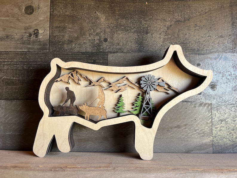 May include: A wooden cutout of a pig with a layered design. The pig is standing on a wooden surface. The cutout features a scene of a person walking a dog in a mountainous landscape with a windmill in the background.
