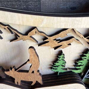 May include: A wooden layered art piece depicting a person riding a deer through a mountain landscape. The deer and person are brown, the mountains are brown, and the trees are green.