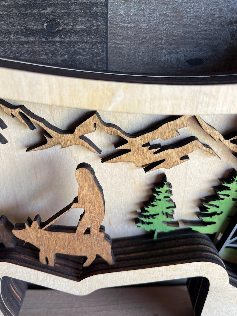 May include: A wooden layered art piece depicting a person riding a deer through a mountain landscape. The deer and person are brown, the mountains are brown, and the trees are green.