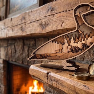 May include: A multi-layered wooden duck silhouette featuring a laser-cut forest landscape with pine trees and a small bird in flight. The sculpture is crafted from light and dark wood tones, creating a depth effect within the duck's body shape.