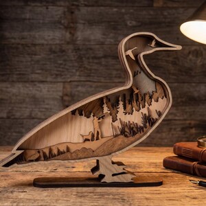 May include: A multi-layered wooden duck silhouette featuring a laser-cut scene of a hunter with a dog in a forest landscape. The interior cutout displays pine trees, tall grass, and flying ducks, all crafted from natural wood tones with varying depths.