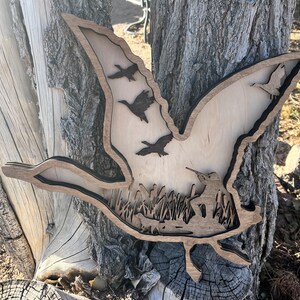 May include: A wooden wall art piece depicting a flying goose with a flock of geese flying behind it. The art piece is cut out of wood and has a natural wood finish. The geese are silhouetted against a background of trees and grass.