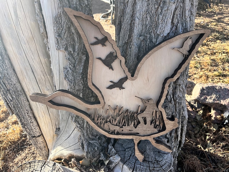 May include: A wooden wall art piece depicting a flying goose with a flock of geese flying behind it. The art piece is cut out of wood and has a natural wood finish. The geese are silhouetted against a background of trees and grass.