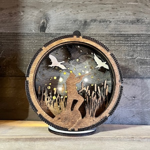 May include: A wooden, circular, laser-cut lamp with a silhouette of a hunter in a field of tall grass and flying ducks. The lamp has a dark wood finish and a white base.