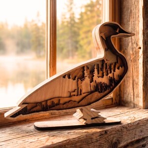 May include: A layered wooden duck sculpture featuring a cutout silhouette of a hunter with a dog in a forest setting. The piece is constructed from multiple stacked wood panels with natural grain patterns, creating a three-dimensional effect.
