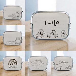 Könnte beinhalten: Eine Auswahl an Edelstahl-Lunchboxen mit verschiedenen Gravuren. Jede Box zeigt eine andere Illustration, darunter ein Bär, ein Monster, ein Regenbogen, ein Dinosaurier und andere Cartoon-Figuren, zusammen mit personalisierten Namen.