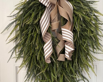 May include: A green, teardrop-shaped wreath with long, slender leaves. The wreath is adorned with a large bow made of striped and solid brown burlap ribbon. The wreath is hanging on a white door.