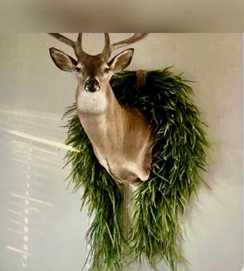 K&ouml;nnte beinhalten: Ein pr&auml;parierter Hirschkopf an der Wand, geschm&uuml;ckt mit einem gr&uuml;nen, blattreichen Kranz. Der Hirsch hat braunes Fell, ein Geweih und eine detaillierte Gesichtsstruktur. Der Kranz ist ein leuchtendes Gr&uuml;n und bildet einen auff&auml;lligen Kontrast zum Fell des Hirsches.