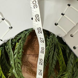 K&ouml;nnte beinhalten: Ein grauer, unfertiger Kranzrohling mit wei&szlig;er Schn&uuml;rung und einer Juteschnur zum Aufh&auml;ngen. Der Kranz ist von gr&uuml;nem Kunstblatt umgeben. Ein Ma&szlig;band zeigt, dass der Kranz etwa 76 cm Durchmesser hat.