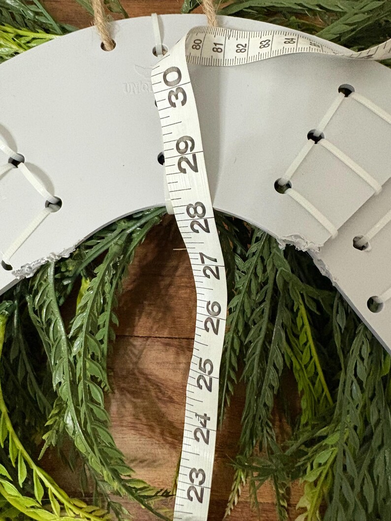 K&ouml;nnte beinhalten: Ein grauer, unfertiger Kranzrohling mit wei&szlig;er Schn&uuml;rung und einer Juteschnur zum Aufh&auml;ngen. Der Kranz ist von gr&uuml;nem Kunstblatt umgeben. Ein Ma&szlig;band zeigt, dass der Kranz etwa 76 cm Durchmesser hat.