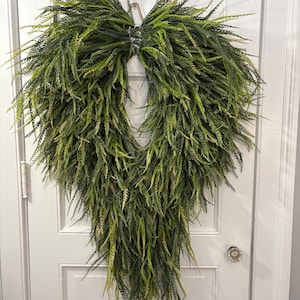 May include: A large, heart-shaped decorative wreath constructed from long, slender, pointed green leaves in varying shades of lime, olive, and dark forest green. The foliage is arranged in a cascading, downward-pointing teardrop formation.