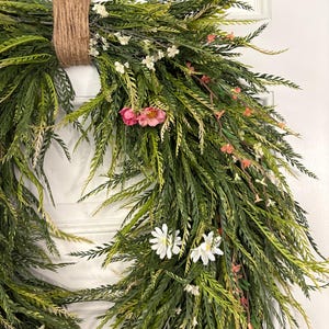 K&ouml;nnte beinhalten: Ein dekorativer Kranz mit &uuml;ppigem, gr&uuml;nem Laub, akzentuiert mit kleinen wei&szlig;en und rosa Bl&uuml;ten. Der Kranz ist mit einer nat&uuml;rlichen Schnurschlaufe zum Aufh&auml;ngen gebunden. Das Design erinnert an eine frische, fr&uuml;hlingshafte &Auml;sthetik, geeignet f&uuml;r T&uuml;r- oder Wanddekoration.