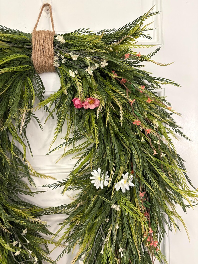 K&ouml;nnte beinhalten: Ein dekorativer Kranz mit &uuml;ppigem, gr&uuml;nem Laub, akzentuiert mit kleinen wei&szlig;en und rosa Bl&uuml;ten. Der Kranz ist mit einer nat&uuml;rlichen Schnurschlaufe zum Aufh&auml;ngen gebunden. Das Design erinnert an eine frische, fr&uuml;hlingshafte &Auml;sthetik, geeignet f&uuml;r T&uuml;r- oder Wanddekoration.