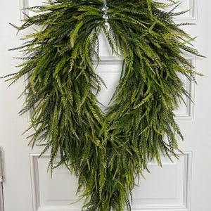 May include: A green and yellow artificial wreath with a teardrop shape. The wreath is made of long, thin, feathery leaves, creating a full and textured appearance. It is hanging on a white door.