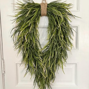 May include: A green and white artificial leaf wreath. The wreath is in an oval shape and is made of long, thin leaves. The leaves are a mix of green and white. The wreath is tied together with a brown twine.
