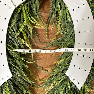 K&ouml;nnte beinhalten: Ein wei&szlig;er hufeisenf&ouml;rmiger Kranz mit einer gr&uuml;n-gelben Blattbasis. Der Kranz ist etwa 35,5 cm lang. Der wei&szlig;e Rahmen hat gleichm&auml;&szlig;ig verteilte L&ouml;cher mit wei&szlig;en N&auml;hten. Der Kranz befindet sich auf einer Holzoberfl&auml;che.
