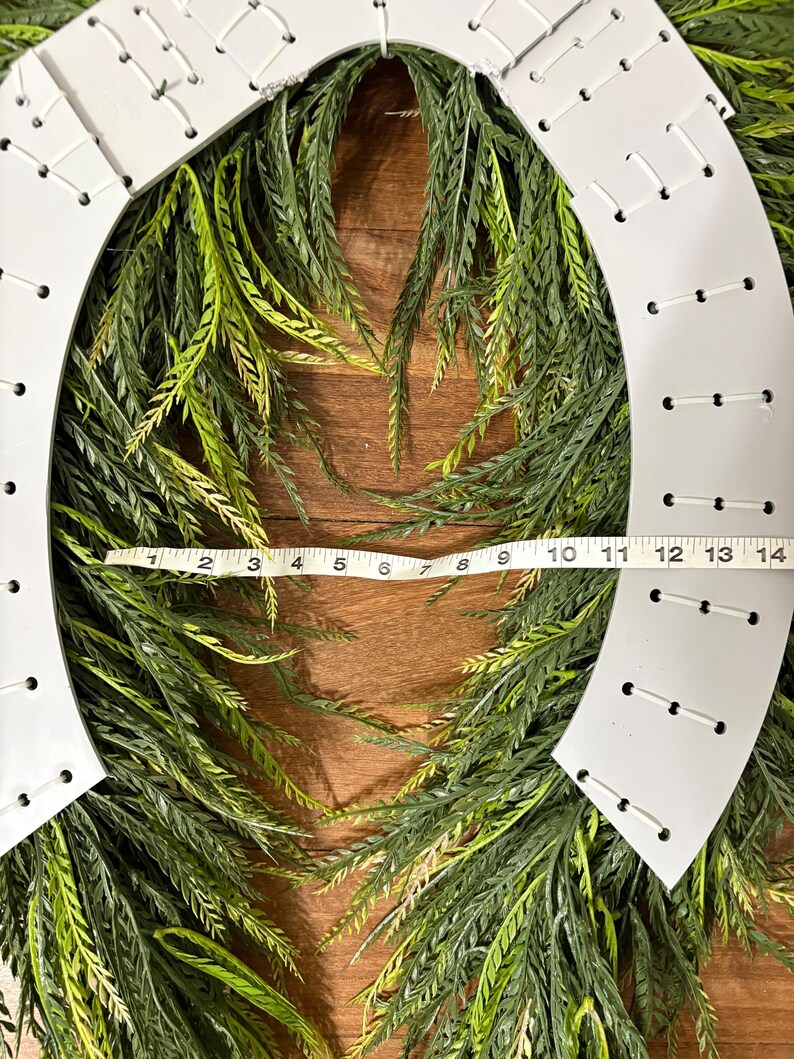K&ouml;nnte beinhalten: Ein wei&szlig;er hufeisenf&ouml;rmiger Kranz mit einer gr&uuml;n-gelben Blattbasis. Der Kranz ist etwa 35,5 cm lang. Der wei&szlig;e Rahmen hat gleichm&auml;&szlig;ig verteilte L&ouml;cher mit wei&szlig;en N&auml;hten. Der Kranz befindet sich auf einer Holzoberfl&auml;che.