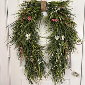 K&ouml;nnte beinhalten: Ein dekorativer Kranz mit &uuml;ppigem gr&uuml;nen Laub, akzentuiert mit kleinen rosa und wei&szlig;en Bl&uuml;ten. Der Kranz wird von einer nat&uuml;rlichen Schnurschlaufe gehalten, ideal, um jedem T&uuml;r oder Wand einen Hauch von Fr&uuml;hling zu verleihen.