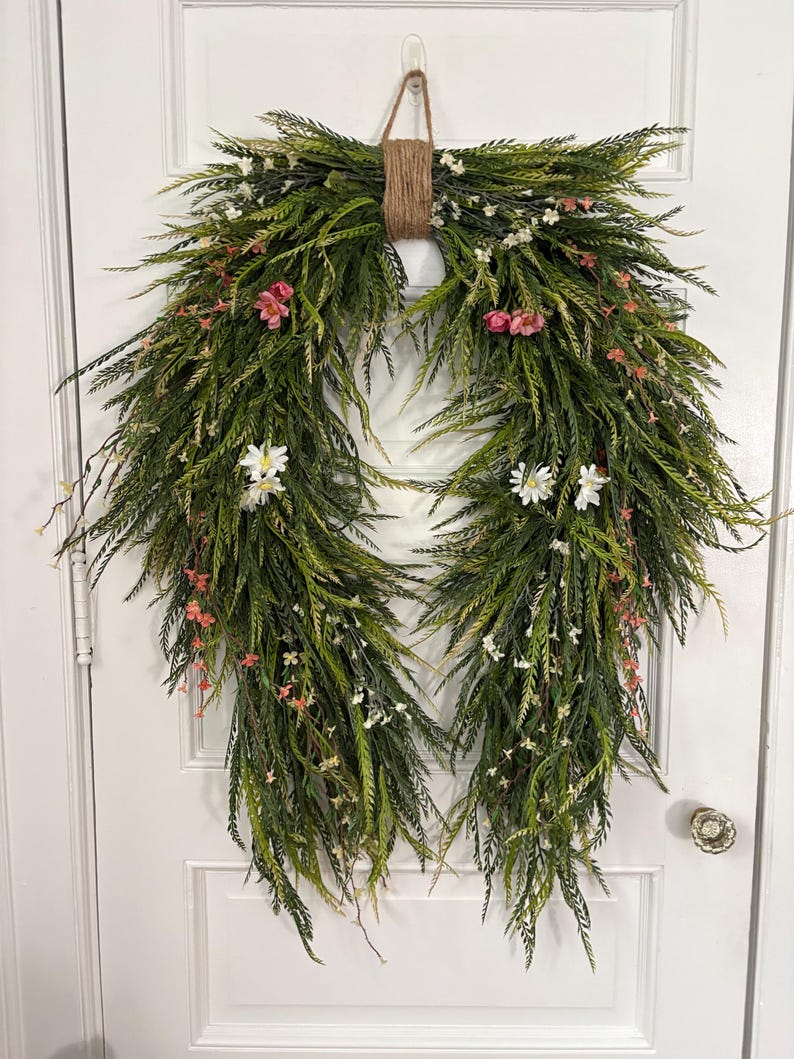 K&ouml;nnte beinhalten: Ein dekorativer Kranz mit &uuml;ppigem gr&uuml;nen Laub, akzentuiert mit kleinen rosa und wei&szlig;en Bl&uuml;ten. Der Kranz wird von einer nat&uuml;rlichen Schnurschlaufe gehalten, ideal, um jedem T&uuml;r oder Wand einen Hauch von Fr&uuml;hling zu verleihen.