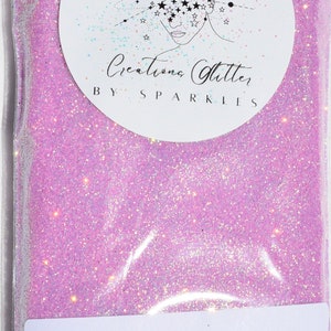 May include: A resealable plastic bag filled with pink glitter with iridescent shimmer. The bag has a white label with the text "Creations Glitter by Sparkles" and a second label with the text "Pink Raindrop 2 oz of the most FABULOUS GLITTER EVER!"