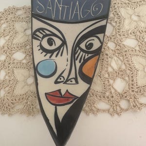 May include: A ceramic decorative piece with a stylized face design. The piece is triangular, with a blue top section bearing the name "SANTIAGO". The face features large eyes, red lips, and blue and orange cheek accents. The background is a cream color.