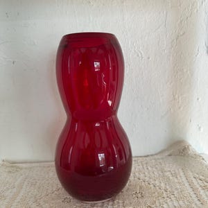 May include: A tall, ruby-red glass vase with a unique hourglass shape. The vase has a wide base that tapers in the middle and flares out again at the top. The glossy surface reflects light, creating highlights and shadows.