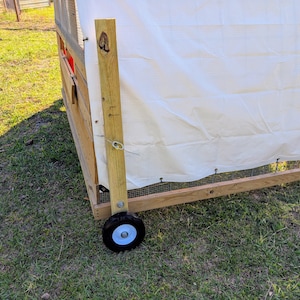 May include: A wooden chicken coop with a white canvas cover and a wheel on the bottom. The coop is on a grassy surface.