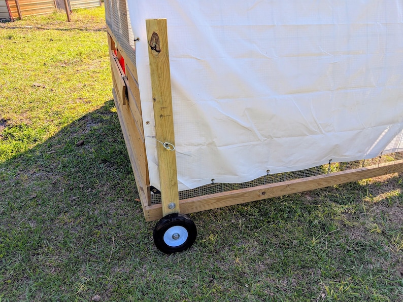 May include: A wooden chicken coop with a white canvas cover and a wheel on the bottom. The coop is on a grassy surface.