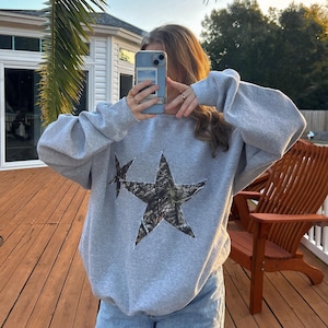 May include: A gray sweatshirt with two large stars on the front. The stars are made of a camouflage fabric.