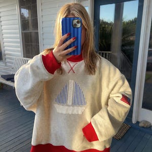 May include: An oatmeal-colored long-sleeve sweatshirt featuring a red mock neck collar, red sleeve cuffs, and a red hem. The front displays a sailboat applique with blue and white striped sails. A small American flag patch is on the left sleeve.