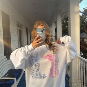 May include: White oversized sweatshirt with a blue and pink floral print of cowboy boots. The sweatshirt has a crew neck and long sleeves.