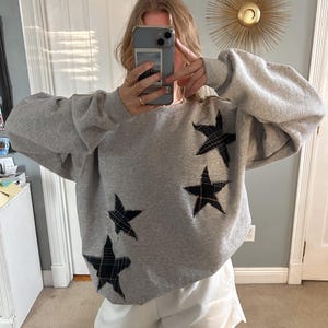 May include: A gray sweatshirt with four black and yellow plaid stars on the front. The sweatshirt is oversized and has a relaxed fit.