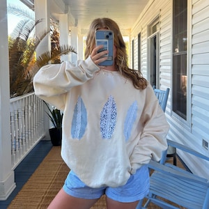 May include: A cream-colored sweatshirt with three blue and white lace appliques in the shape of leaves. The sweatshirt is oversized and has a relaxed fit. The model is wearing blue and white striped shorts.