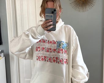 May include: White hoodie with a floral print resembling an American flag. The print features pink, red, and blue flowers with white lace accents.