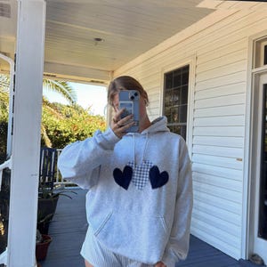 May include: A gray hoodie with a black and white checkered heart design. The hoodie is worn by a person with short brown hair. The person is standing on a wooden porch.
