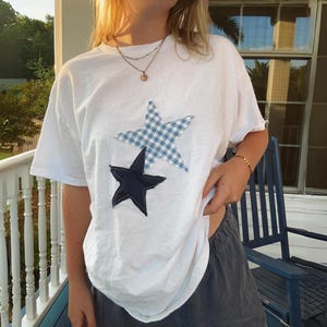 May include: White t-shirt with two fabric star appliques. The larger star is blue and white gingham, and the smaller star is navy blue.