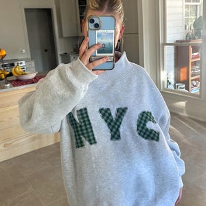 May include: A light gray sweatshirt with the letters "NYC" appliqued in green plaid fabric. The sweatshirt has a high neck and long sleeves. The person is holding a phone with a picture on the screen.