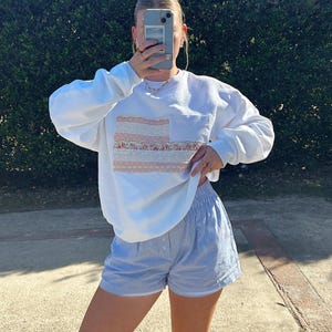 May include: A white sweatshirt with a pink and white floral design on the front pocket. The sweatshirt is paired with blue and white striped shorts.