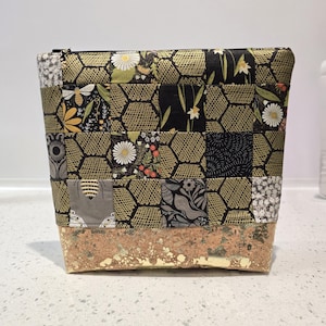 May include: A patchwork cosmetic bag with a gold honeycomb pattern and floral fabric panels. The base is a textured, light brown material. The bag has a black zipper closure.