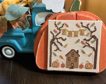 Könnte beinhalten: Ein Kreuzstichmuster einer kleinen Stadt mit einem Haus, Bäumen und Kürbissen, gerahmt in einem hölzernen Kürbis. Das Muster trägt den Titel "Fall in a Small Town" und ist ein Originaldesign von Small Town Needleworks.