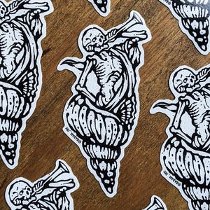 Può includere: Una collezione di adesivi bianchi con un'illustrazione nera di uno scheletro che suona la tromba mentre emerge da una conchiglia. Gli adesivi sono su una superficie di legno. Il nome dell'artista, @BADAM_INK, è stampato su ogni adesivo.