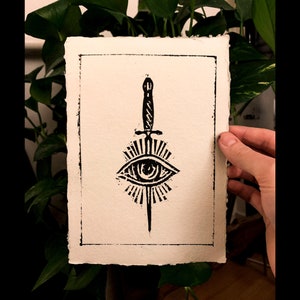 May include: Black and white print of a stylized eye with a dagger piercing through it. The eye is surrounded by a radiating pattern of lines.