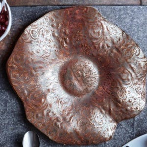 Puede incluir: Un plato decorativo de color cobre con un patrón floral en relieve. El plato tiene una zona central ligeramente elevada y una forma irregular y orgánica. El acabado metálico le da un aspecto rústico y artesanal.